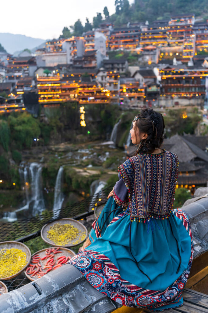 Atardecer en la antigua ciudad de Lijiang iluminada con luces cálidas, mientras una mujer china observa el paisaje tradicional.